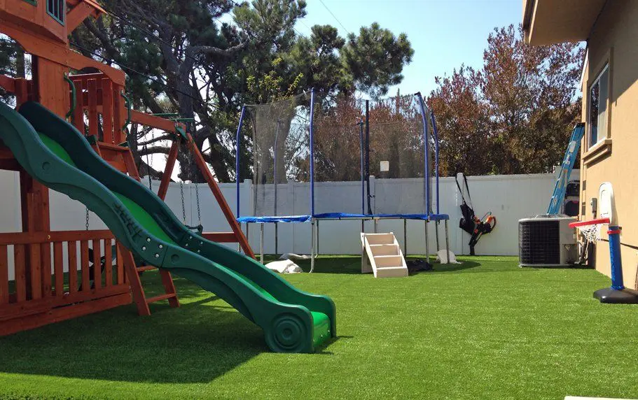 A backyard playground with a green slide attached to a wooden play structure, a trampoline with a safety net, a swing set, and a basketball hoop with a small backboard. The ground is meticulously covered in artificial grass landscaping, and a white fence encloses the yard.
