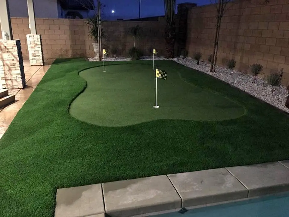 A small putting green with three holes, each marked by a yellow checkered flag, in a backyard surrounded by landscaping rocks and a brick wall. The neatly trimmed artificial grass landscaping is expertly installed, and the scene is lit in the evening. Contact us for free estimates on your project!